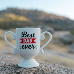 Tienda A white trophy-shaped mug with 'Best Dad Ever' text placed on a rock in a natural outdoor setting.